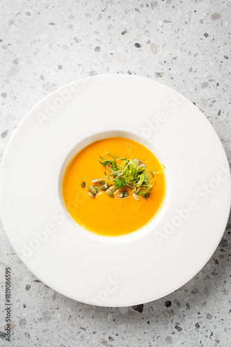 A close-up shot of vibrant orange pumpkin soup, elegantly garnished with green sprouts and pumpkin seeds, served in a white bowl. Appetizing and healthy dish, top view