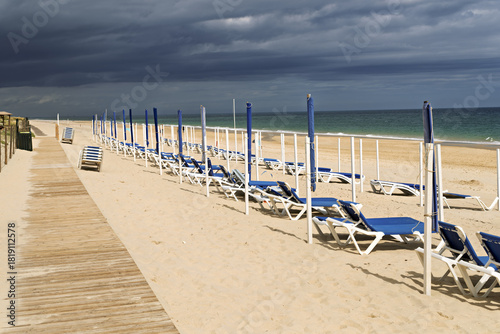 Beach scene with rows of empty blue lounge chairs and white-and-blue umbrellas neatly arranged on golden sand on Ilha de Tavira, Portugal,