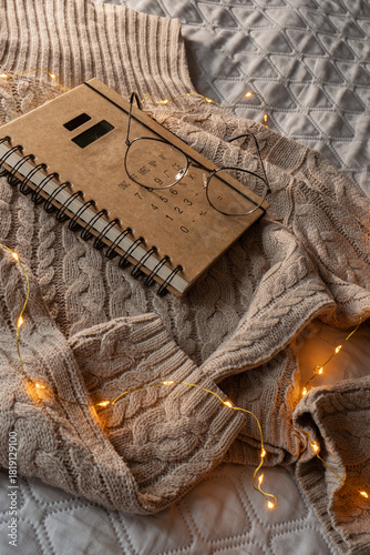 A beige warm sweater on a bad with glasses and book. Cozy home background with glowing Christmas lights