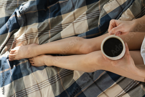 Woman holding a cup of coffee drink on legs and sitting down on a bed. Cozy home and beauty concept