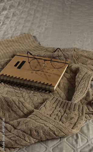 A beige warm sweater on a bad with glasses and book. Cozy home background