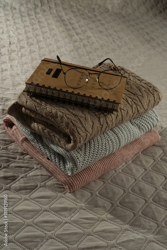 Stack of warm sweaters, book and glasses on a bed