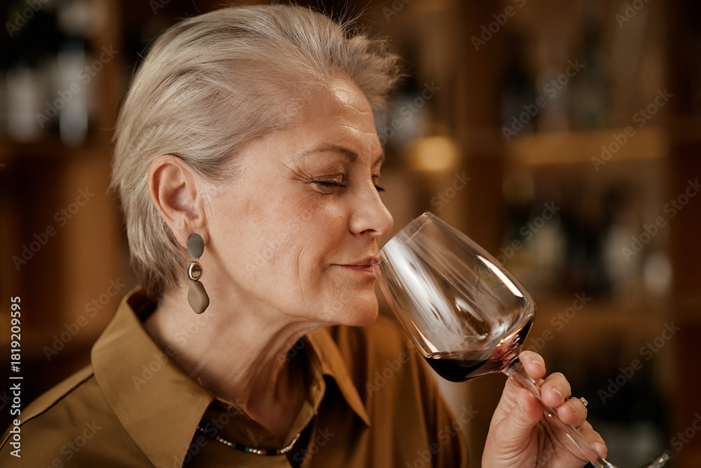 Naklejka premium Senior Caucasian woman holding wine glass, smelling red wine during tasting session, eyes closed, short gray hair, standing in front of blurred wine bottles on shelves