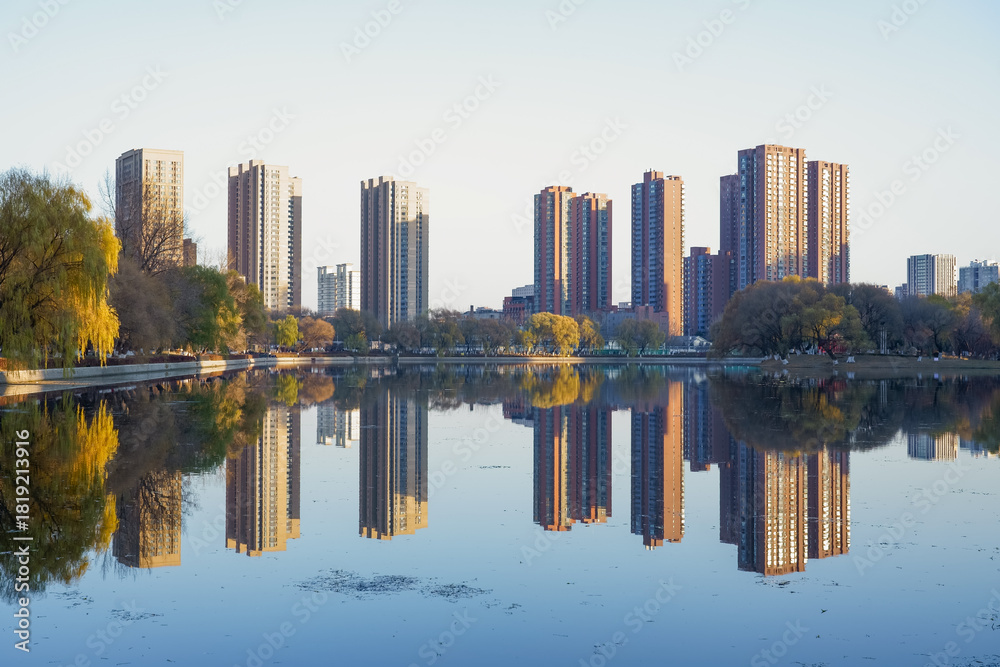 Obraz premium Calm waters of Diaoyu Lake perfectly mirror the modern buildings of Shenyang, creating a serene urban landscape. Soft daylight enhances the symmetry and clarity of the reflection.