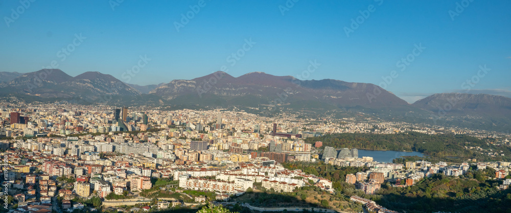 Naklejka premium A warm, sweeping panorama of Tirana's expansive cityscape and the Artificial Lake, set against the majestic Dajti Mountain range under the late afternoon light.