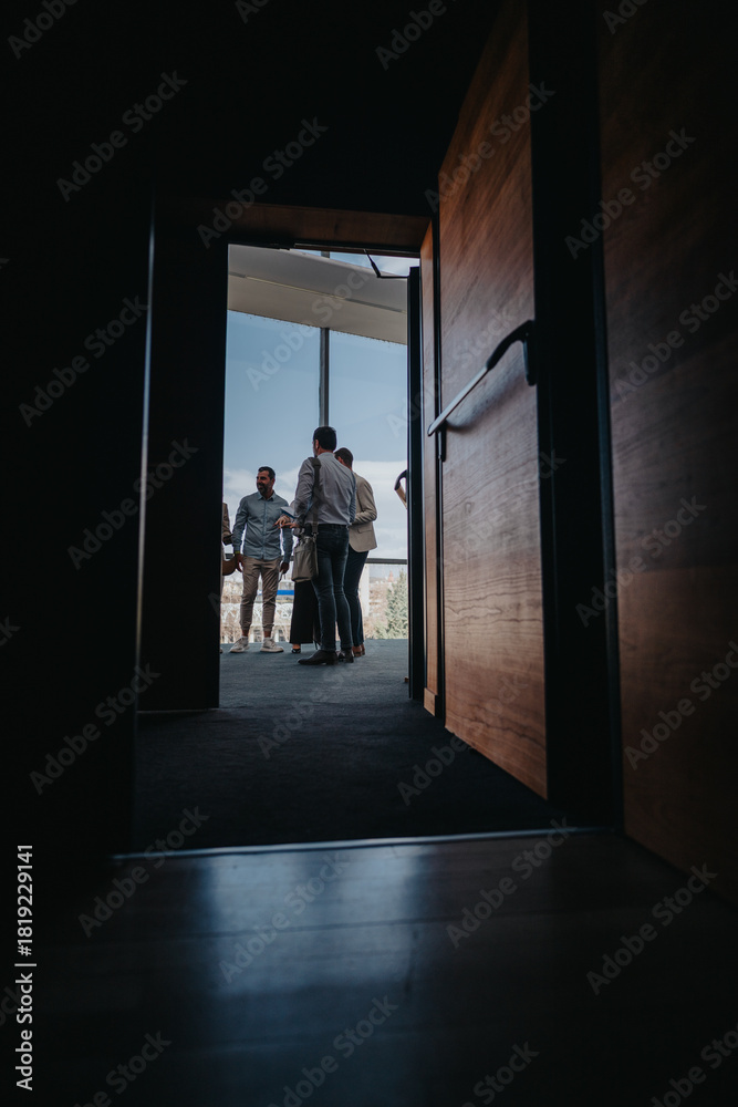 Obraz premium A dark hallway opens to a bright terrace where colleagues stand and converse near large windows. The scene centers on people sharing a casual moment, suggesting connection during a break.