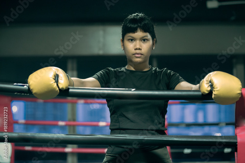 Intense Asian Female Boxer with Golden Gloves Leaning on Ring Ropes indoors night time