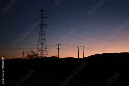 High Country Salmon area, New Zealand - May 23, 2025: A dramatic silhouette of power lines and transmission towers against a mountain backdrop during twilight. The sky transitions from orange to blue 