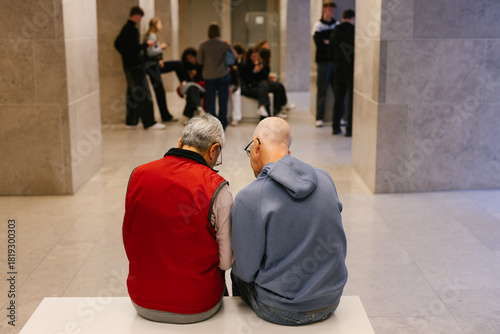 Two older people, a couple, sit on a bench in a museum.