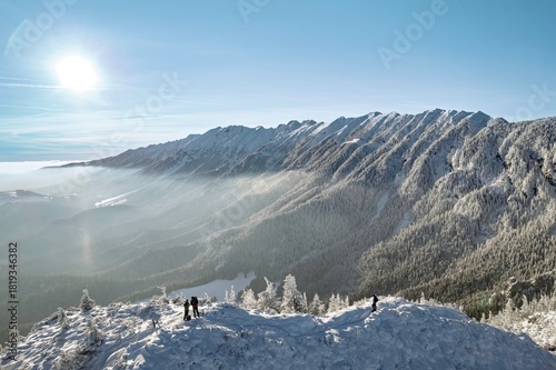 Adventurers stand atop a snowy peak, gazing at the Piatra Craiului and Bucegi Mountains under a bright sun. The winter landscape showcases frosted trees and a serene atmosphere