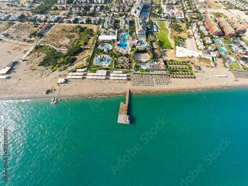 Fototapeta Naklejka Na Ścianę i Meble -  Aerial view of a wide sandy beach, turquoise sea, and a wooden pier extending out from a row of beachfront resort facilities and sun loungers
