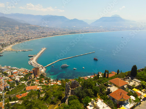 Wide aerial view showing the Alanya harbor pier, the Red Tower (Kızıl Kule) area, ships in the turquoise bay, and the extensive city stretching along the coast