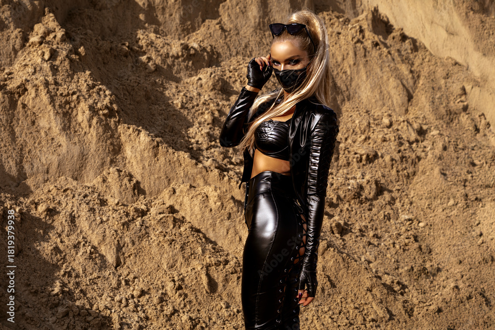 Obraz premium Futuristic woman in black leather outfit posing confidently in desert sand landscape with dynamic hair movement