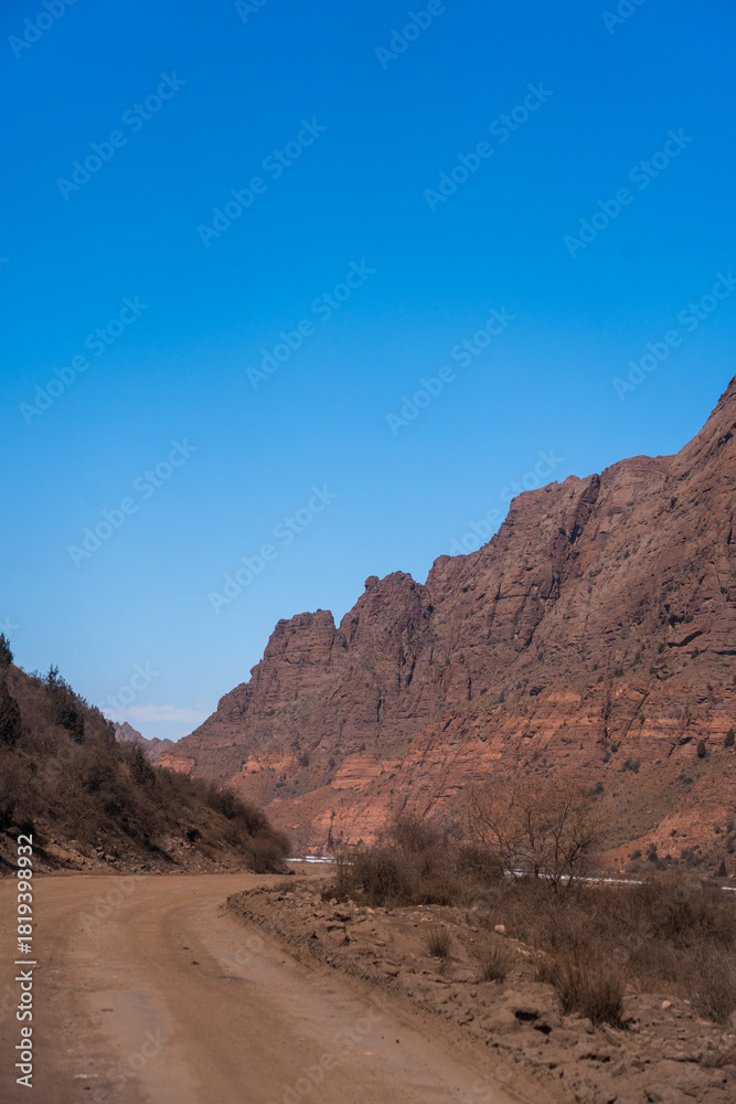 Fototapeta premium An unpaved road winding through a tight mountain pass or valley framed by massive, rugged red rock cliffs under a bright blue sky