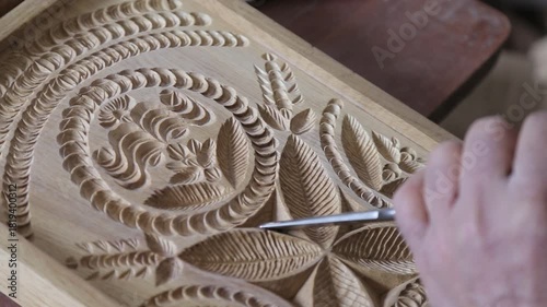Close-up of an artisan's hand using a chisel to carve intricate floral ornaments into a wooden board, showcasing traditional woodworking skills and precise attention to beautiful artistic details.

