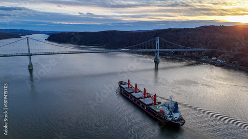 Aerial view of a colossal cargo ship glides under the Mid-Hudson Bridge, connecting the tranquil waters under a painted sky, Poughkeepsie, New York, United States.