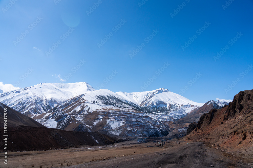Fototapeta premium Wide panoramic view of a vast landscape featuring snow-capped mountains and a large, visible open-pit mine site on the lower slopes