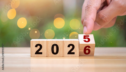 Wooden blocks displaying transition from 2025 to 2026, with hand turning block. background features bokeh effect with warm lights, creating festive and hopeful atmosphere
