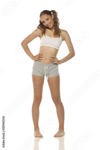 Studio shot of young woman in white top and grey shorts smiling with hands on hips on white background, full body