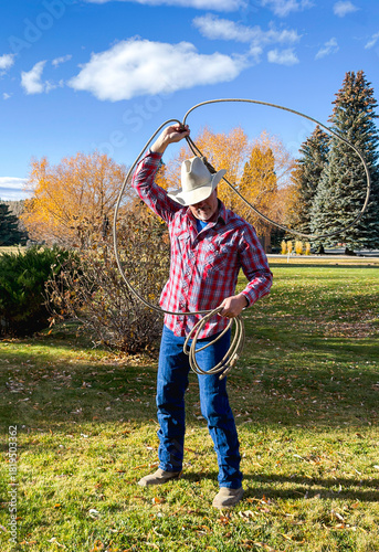 Western rodeo cowboy swings rope  above  head with lariat in his hand. He wears typical cowboy hat, red plaid shirt, jeans & working boots. Setting is park tree yard in the fall, Wyoming, USA..