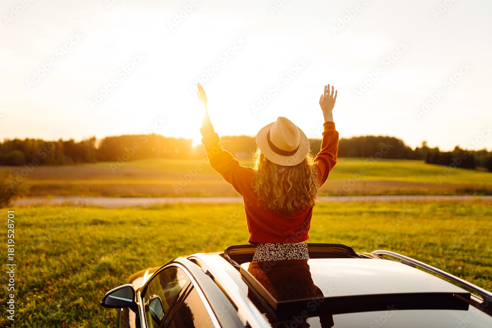 Fototapeta premium A joyful woman leans out of a car window on a sunny day at sunset. A young woman in a hat enjoys nature and feels the freedom of a road trip. Concept of adventure and relaxation.