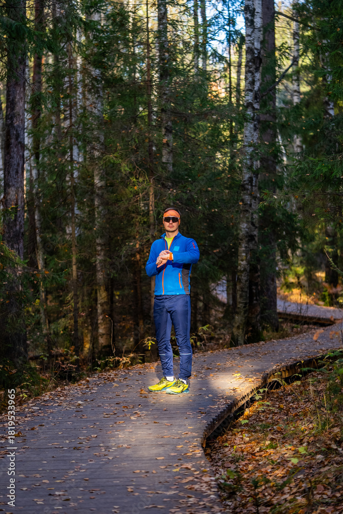 Obraz premium Man on winding wooden trail in autumn park checks his smartwatch before a run, surrounded by evergreen trees and fallen leaves, focused on fitness and training
