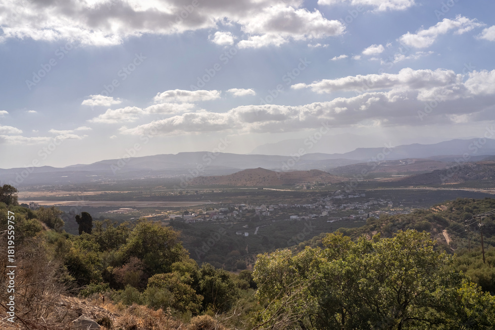 Obraz premium Wide panoramic view of a sunlit Cretan valley with distant mountains, soft clouds, and rays of light breaking through the sky. The scene overlooks a small village surrounded by olive groves
