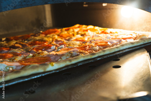 Large rectangular artisan pizza placed on a peel inside a professional electric oven. Freshly baked crust with visible texture, captured in a warm kitchen environment. Concept of bakery product
