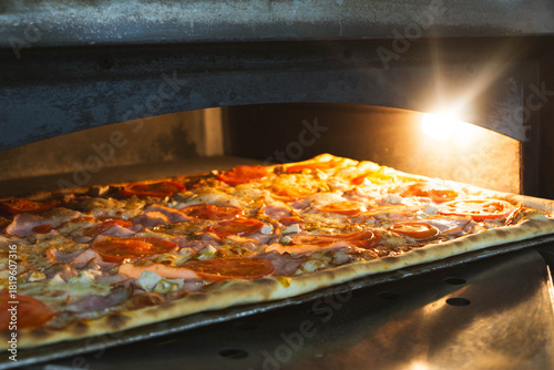 Large rectangular artisan pizza placed on a peel inside a professional electric oven. Freshly baked crust with visible texture, captured in a warm kitchen environment. Concept of bakery product