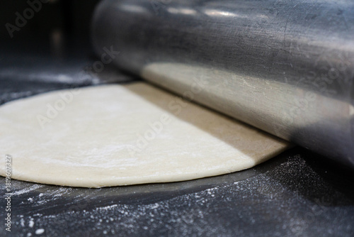 Close-up of pizza dough being rolled out with a large heavy-duty steel rolling pin on a clean stainless steel worktable.Smooth dough texture, metallic reflections, and authentic cooking process.