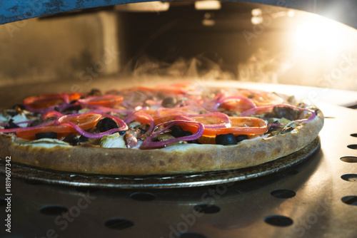 Hot freshly baked small pizza resting on a steel peel inside a professional electric oven. Crispy golden crust with toppings including mushrooms, olives, tomatoes, sausages, chicken, and onions