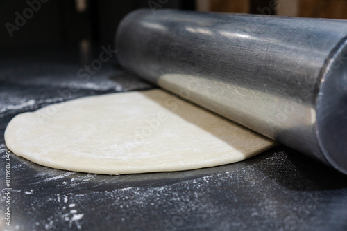 Close-up of pizza dough being rolled out with a large heavy-duty steel rolling pin on a clean stainless steel worktable.Smooth dough texture, metallic reflections, and authentic cooking process.