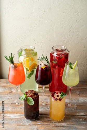 A vibrant display of various drinks sits on a rustic wooden table. Each glass is adorned with fresh fruit, herbs, and colorful layers, inviting enjoyment on a sunny day
