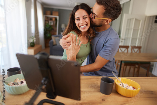 A young couple enjoys a cozy meal together at home, smiling and showing off an engagement ring while connecting with loved ones through a video call.