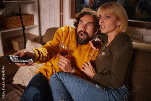 A couple sits closely on a sofa, engrossed in a tense movie scene. They hold glasses of wine, showing excitement and surprise as they watch the screen.