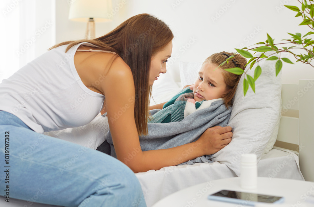Fototapeta premium Motherhood, health and support. Young caring mother comforts and cares for her son who looks upset and unhealthy. In bright bedroom, child is wrapped in soft blanket, looking for warmth and comfort.