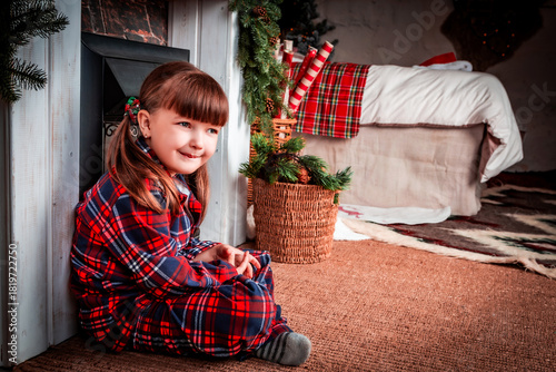 Happy little girl child in pajamas sitting in christmas decorated room near fireplace. Christmas. New Year. St. Nicholas Day. Cozy, festive bedroom interior.	