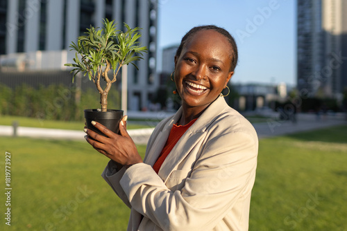 Young African woman holds potted plant in urban park during sunny afternoon