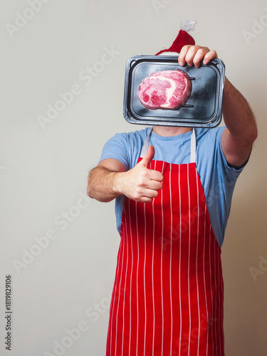 Fine rib eye steak presentation by a male in his 50s in blue shirt and white and red stripe classic butcher apron and Santa Claus hat. High quality beef meat marketing and promotion.