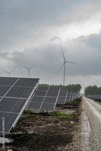 Solar farm with wind turbines in the background on a cloudy day. Renewable energy infrastructure combining solar and wind power in a sustainable energy landscape.