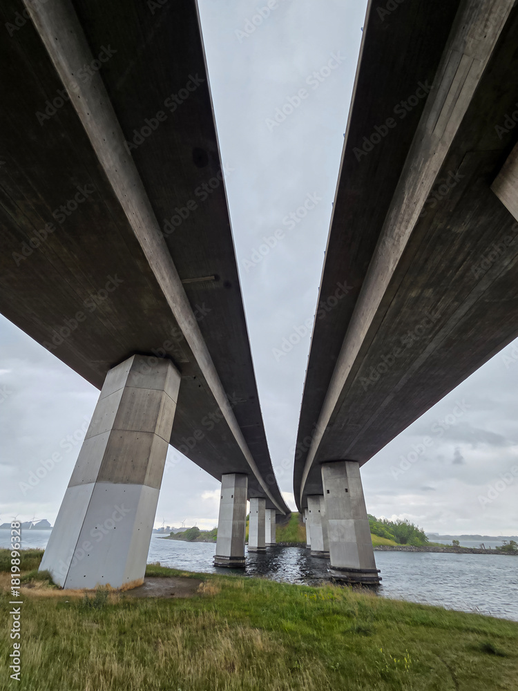 Fototapeta premium A low-angle, centered perspective captures the colossal concrete piers and dual decks of a modern highway bridge spanning a dark, coastal waterway under an overcast sky.