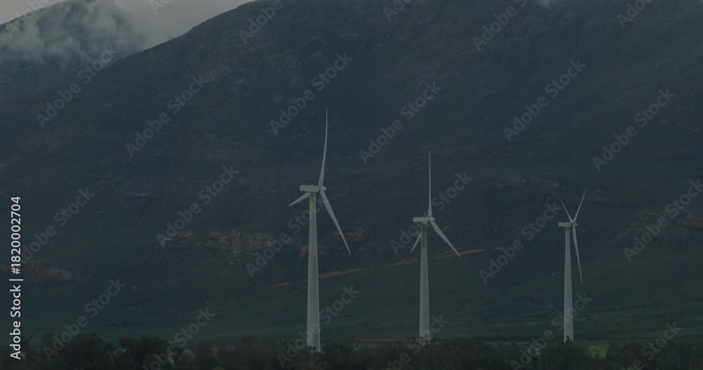Obraz premium Spinning white wind turbines standing at mountain base, with treeline, rocky slopes, mist