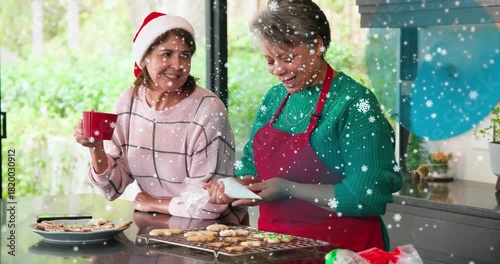 Baker pressing piping bag while daughter sipping red mug, icing holiday cookies with snow overlays