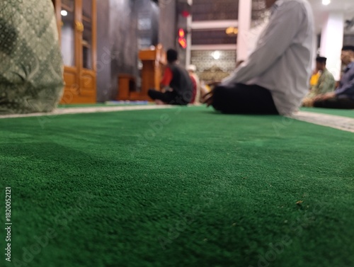 Close up of a vibrant green prayer rug with blurred people praying. Muslim men sitting on a clean green carpet during a worship service. Low angle view of a mosque interior