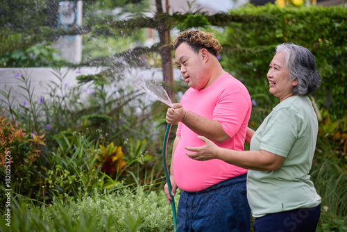 A happy moment of a senior woman and a young man with down syndrome holding a water hose in a garden, enjoying outdoor activity together