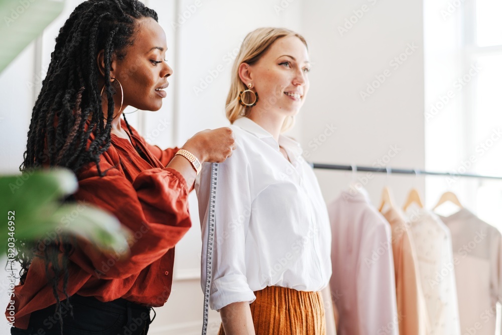Fototapeta premium A Black stylist woman fitting outfit to Caucasian woman model in a stylish clothing store. The Black woman is helping the White woman with fitting, showcasing outfit fashion and personal styling.