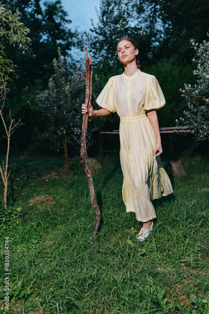 Fototapeta premium A woman in a pale yellow dress walks outdoors in a lush garden, holding a wooden staff, with calm expression and soft natural light, surrounding greenery creating a tranquil serene atmosphere