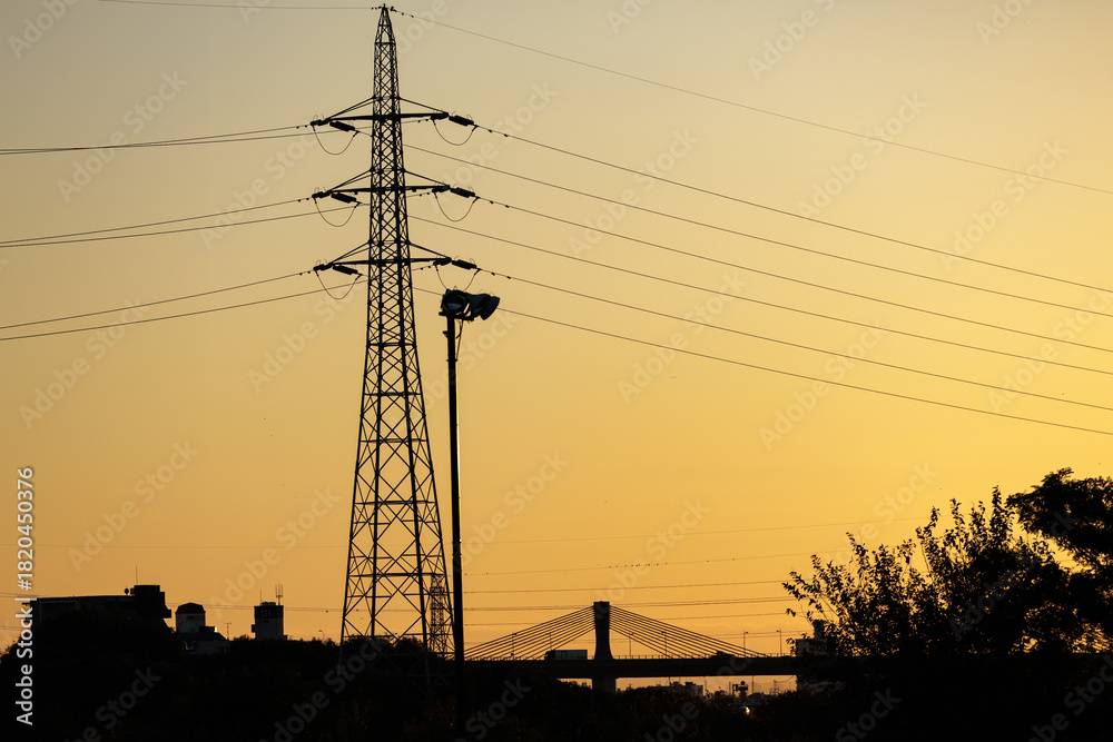 Fototapeta premium Golden sunset silhouettes landscape of high voltage tower and taut power lines towering above trucks crossing cable-stayed suspension bridge.