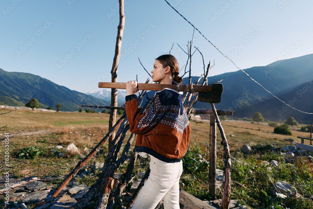 Fototapeta premium Woman carrying wood on rustic fence in mountains, wearing a warm sweater in a rural landscape; portrait of outdoor activity and traditional lifestyle on a sunny day.