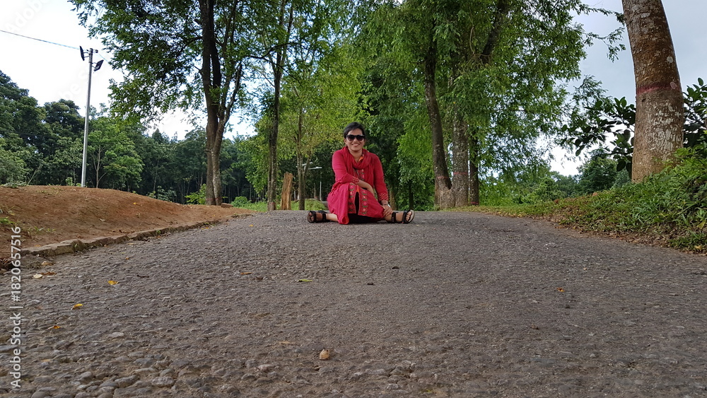 Fototapeta premium Woman in Red Outfit Sitting on Road Surrounded by Trees on a Sunny Day, Nature Portrait